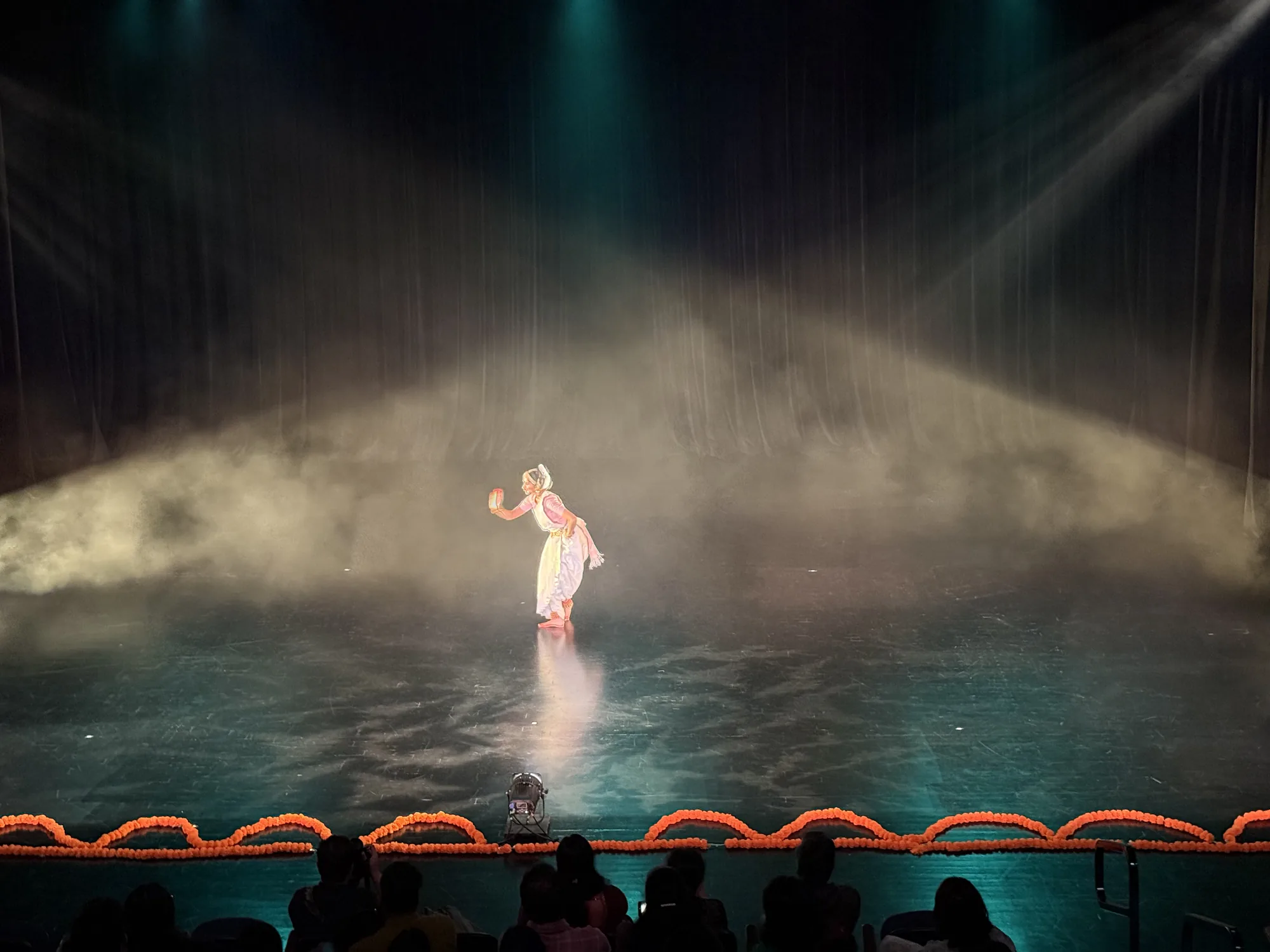 Indian classical dancer performing on a stage in white light and haze, with audience silhouettes in the foreground